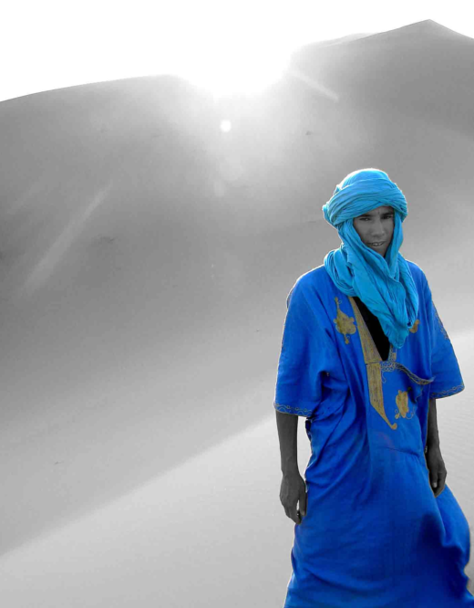 A Bedouin in the dessert wearing blue traditional clothing with the rest of the picture on black and white