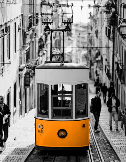 A car of the famous 28th tram line in Lisbon, climbing a steep street. The front of the car is yellow and the rest of the picture is black and white